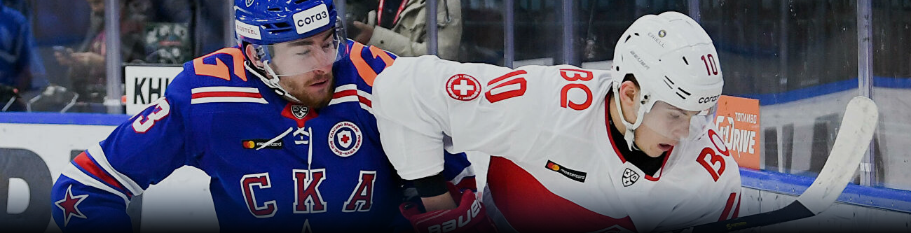 Хоккеисты в синей и белой/красной форме борются за шайбу у борта на арене. Игрок в синем пытается удержать соперника, держа его за плечо. На заднем плане видны трибуны и защитное стекло