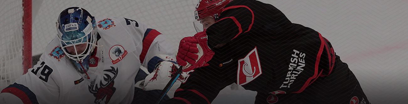 На изображении два хоккеиста во время игры: один в белой форме с синей и красной отделкой, второй в чёрной форме с элементами красного. Игрок в белом наклонился, а игрок в чёрном атакует с поднятой клюшкой